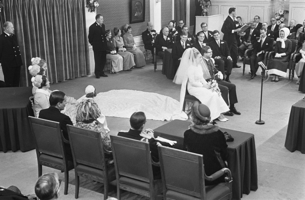 Schwarz-weiß-Hochzeitszeremonie mit sitzenden und stehenden Menschen, Mann mit Mikrofon, Frau mit Blumenstrauß, Podium mit Mikrofon rechts, Blumenschmuck und Vorhang links, gerahmtes Foto und Tür im Hintergrund.