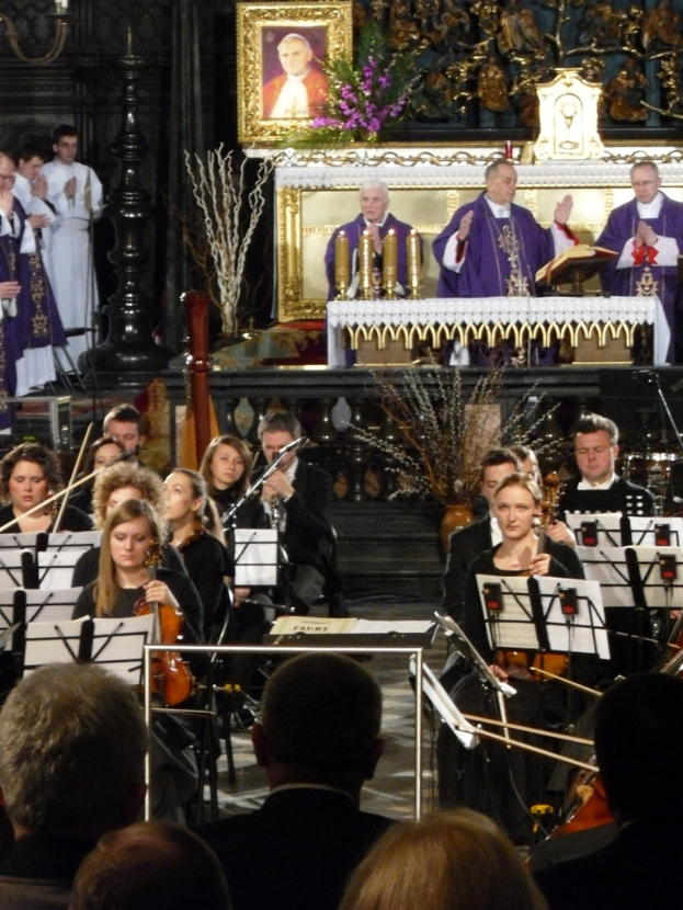 Eine Gruppe von Menschen, die Geigen spielen, mit Notenblättern vor sich, mit drei Kirchenvätern und einer Bibel im Hintergrund und einem Rahmen mit Verzierungen drumherum.