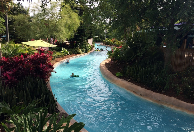 Ein Schwimmbad mit schwimmenden Menschen, umgeben von Pflanzen, Bäumen und einem Zaun, mit Menschen, Sonnenschirmen und Gebäuden im Hintergrund und Pfosten auf beiden Seiten.