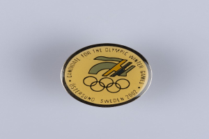 Ein runder Abzeichenaufkleber mit schwarzem Hintergrund und goldfarbenem Olympia-Logo in der Mitte, mit der Beschriftung "Olympische Winterspiele" in fetter weißer Schrift.