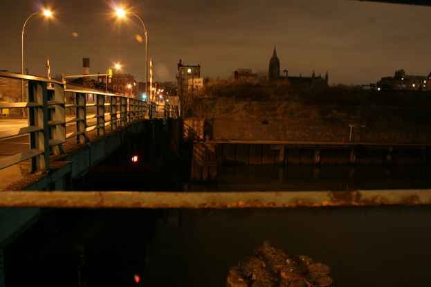 Eine Brücke überspannt einen Fluss mit mehreren Straßenlaternen entlang ihrer Länge und zahlreichen Gebäuden im Hintergrund.