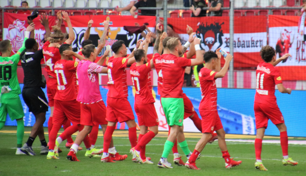 Gruppe von Fußballspielern, die ein Tor feiern, einige halten Flaschen oder klatschen, mit Bannern, einem Zaun, Stühlen und einer Dachkonstruktion im Hintergrund.