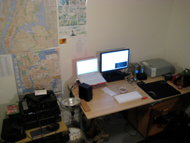Ein Raum mit einem Tisch, auf dem ein Laptop, ein Lautsprecher, ein Monitor, eine Tastatur, ein Ball, eine Maus, ein Scanner und Papiere liegen, und einem weiteren Tisch mit Elektronikgeräten im Hintergrund, mit Charts und Postern an der Wand dahinter.