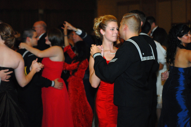 Eine Gruppe elegant gekleideter Menschen tanzt energisch auf einer Tanzfläche mit einer Wand im Hintergrund.