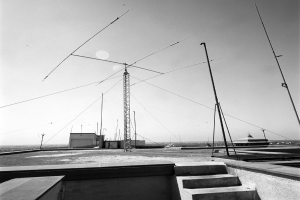 Ein Schwarz-Weiß-Foto einer Funkantenne auf einem Dach, umgeben von Pfählen und Gebäuden, mit Stufen, die zu ihr führen.