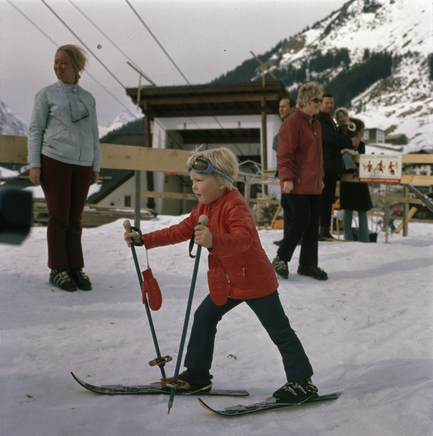 Ein junger Junge in roter Jacke fährt auf einer schneebedeckten Hülle Ski, hält Ski-Stöcke in den Händen, mit Menschen, einem hölzernen Zaun, einer Hütte und einer Tafel mit Schrift im Hintergrund, unter einem sichtbaren Himmel.