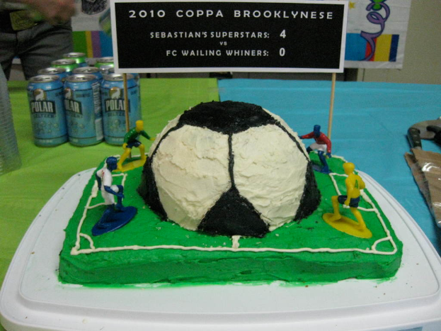 Ein Fußballstadion-geformter Kuchen mit einem Fußball und vier Spielern, umgeben von Softdrink-Dosen, mit einem Banner im Hintergrund und einer Person auf der linken Seite, mit einer Tafel auf dem Kuchen.