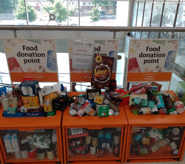 Eine Spendenstation mit orangefarbenen Schränken, die Flaschen, Dosen, Kartons und andere Gegenstände enthalten, begleitet von Informationsschildern, durch Glasfenster betrachtet, die Fahrzeuge, Bäume, Gebäude und Himmel zeigen.