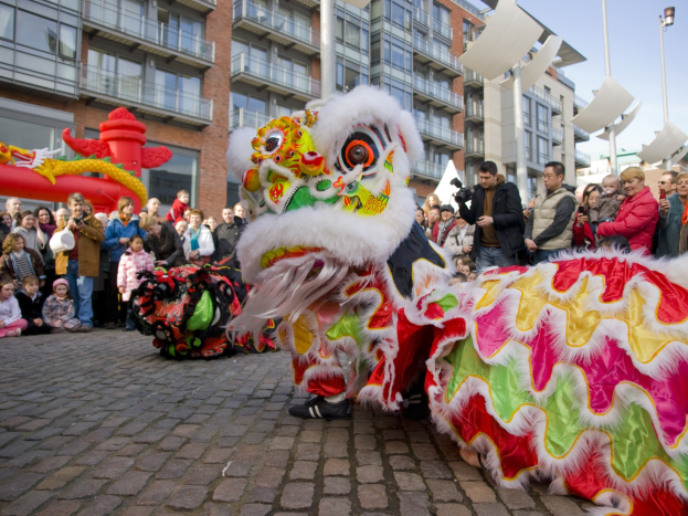 Ein lebendiges chinesisches Neujahrsfest in Amsterdam mit einer Drachenvorführung und einer Menge Schaulustiger, einige fotografieren das Ereignis, vor einem Hintergrund aus Gebäuden, Laternenmästen und einem klaren blauen Himmel.