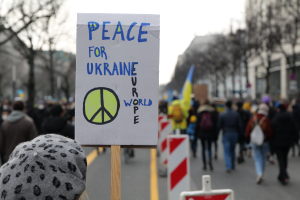 Eine Person mit Mütze hält ein 'Frieden für die Ukraine'-Schild vor einer Menge, die auf einer Straße geht, einige halten Fahnen, mit Bäumen, Gebäuden und einem klaren blauen Himmel im Hintergrund.