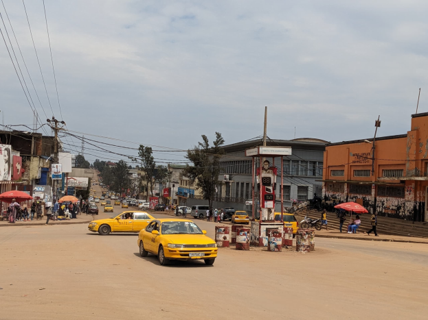 Ein gelbes Taxi fährt eine belebte Stadtstraße mit Gebäuden an den Seiten hinunter, während Fußgänger mit Regenschirmen auf dem Gehweg darunter gehen, unter einem bewölkten Himmel.