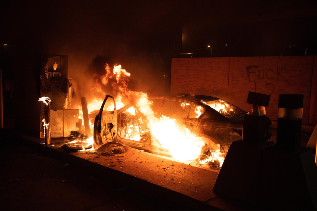 Ein in Flammen stehendes Auto an der Straßense bei Nacht, mit verstreuten Trömmern und einer Wand im Hintergrund.