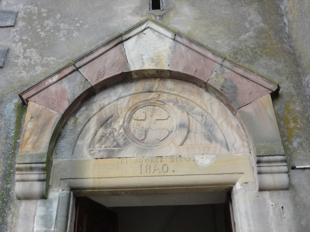 Alter Steinbau mit mittigem gewölbten Eingang, einer verzierten Einlegearbeit darüber und verblasster Text an der Wand.