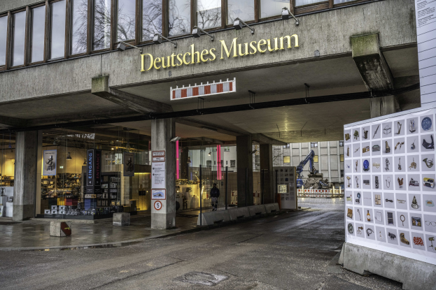 Außenansicht des Deutschen Museums in Berlin, das einen großen Gebäudekomplex mit Glasfenstern, Säulen und einem Namensschild zeigt, sowie ein Informationsdisplay, umgeben von anderen Gebäuden, Bäumen und Himmel.