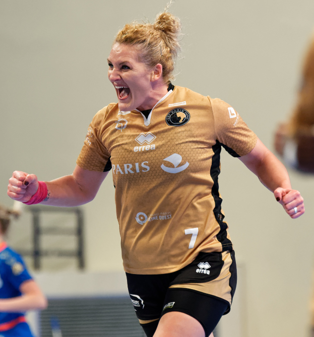 Eine Frau in braunem T-Shirt und schwarzen Shorts rennt auf einem Futsal-Platz, feiert mit triumphaler Miene, während zwei andere Frauen im Hintergrund unscharf sind.