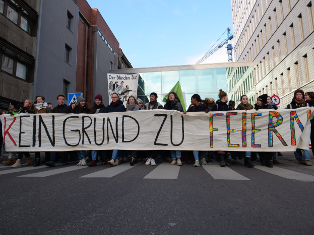Protestierende auf einer Straße halten ein Banner mit der Aufschrift "Kein Grund zu Feiern" gegen deutsche Sparmaßnahmen, mit Gebäuden, einem Schild, einem weiteren Banner, einem Kran und einem bewölkten Himmel im Hintergrund.