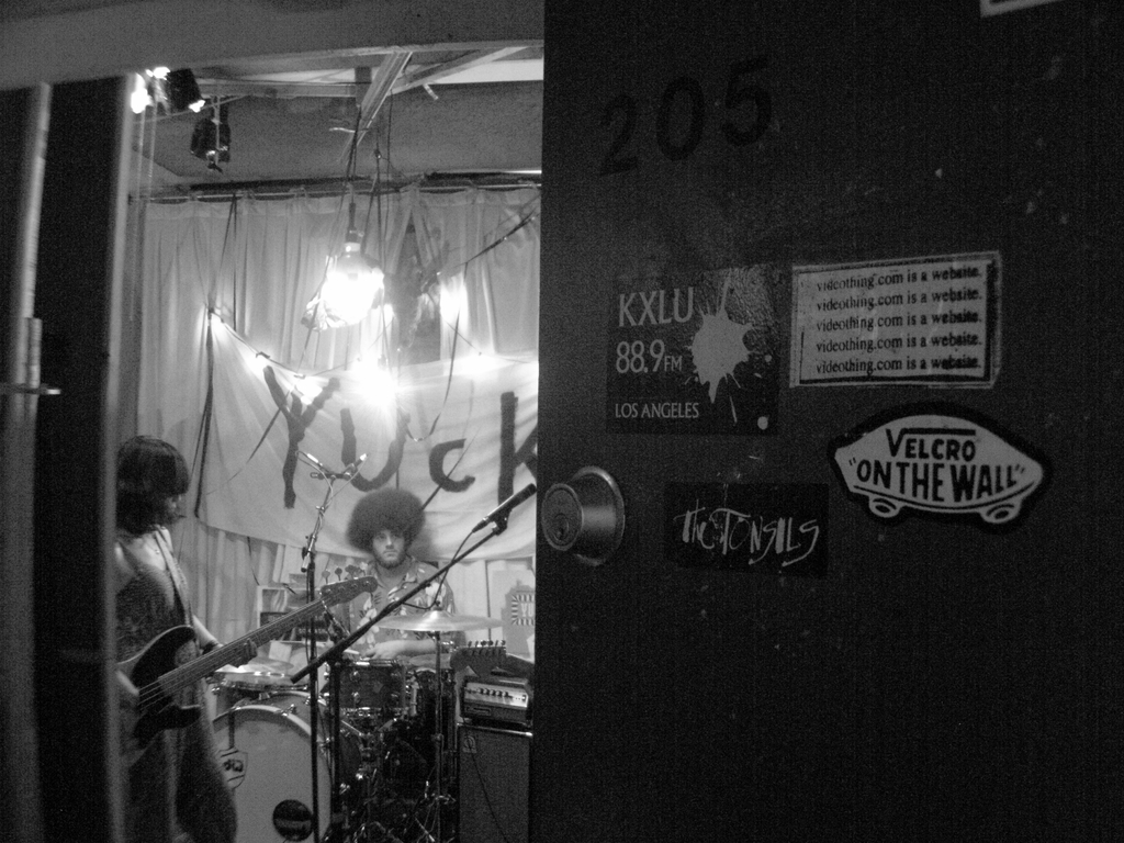 Ein Schwarz-Weiß-Bild eines Mannes, der vor Trommeln sitzt, einer Frau, die Gitarre spielt, und einem Banner mit der Aufschrift 'YUCK' im Hintergrund, sowie einer sichtbaren Tür.