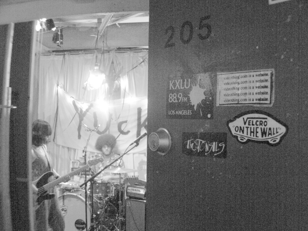 Ein Schwarz-Weiß-Bild eines Mannes, der vor Trommeln sitzt, einer Frau, die Gitarre spielt, und einem Banner mit der Aufschrift 'YUCK' im Hintergrund, sowie einer sichtbaren Tür.