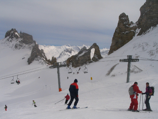 Menschen in Pullovern fahren mit Skiern auf dem Eis, mit einer Seilbahn, Bergen und einem bewölkten Himmel im Hintergrund.