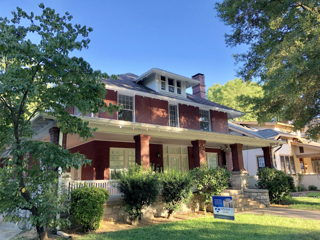Rotes Backsteinhaus mit einem "Immobilien zum Verkauf in Houston, TX"-Schild davor, umgeben von Grün und Stufen zum Eingang.