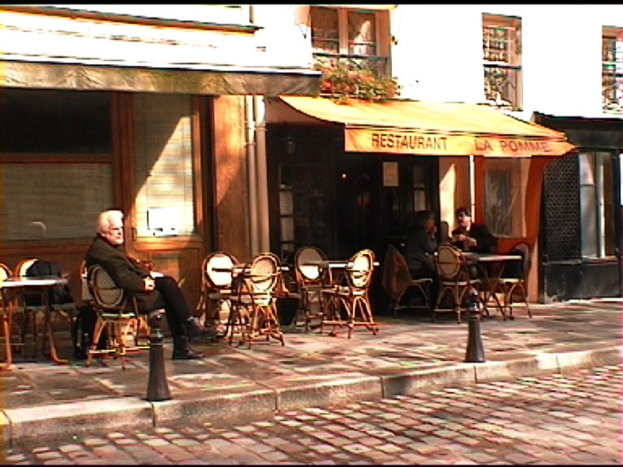 Ein Mann sitzt auf einem Stuhl auf einem Gehweg mit Tischen und Stühlen, vor einem Gebäude mit einer Tafel, auf der "Restaurant" steht.