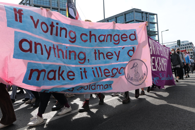 Eine Gruppe von Menschen hält ein Transparent mit der Aufschrift "Wenn Abstimmen etwas ändern würde, hätten sie es verboten", während sie eine Straße mit Gebäuden, Laternenmasten und einem klaren blauen Himmel im Hintergrund entlanggehen.