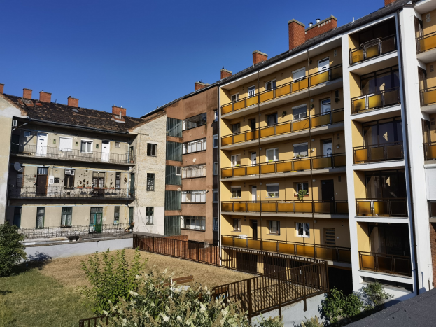 Wohngebäude mit Innenhof, der von grünem Gras, Pflanzen, Bäumen, Fenstern, Türen, Geländern und einem sichtbaren Himmel umgeben ist.