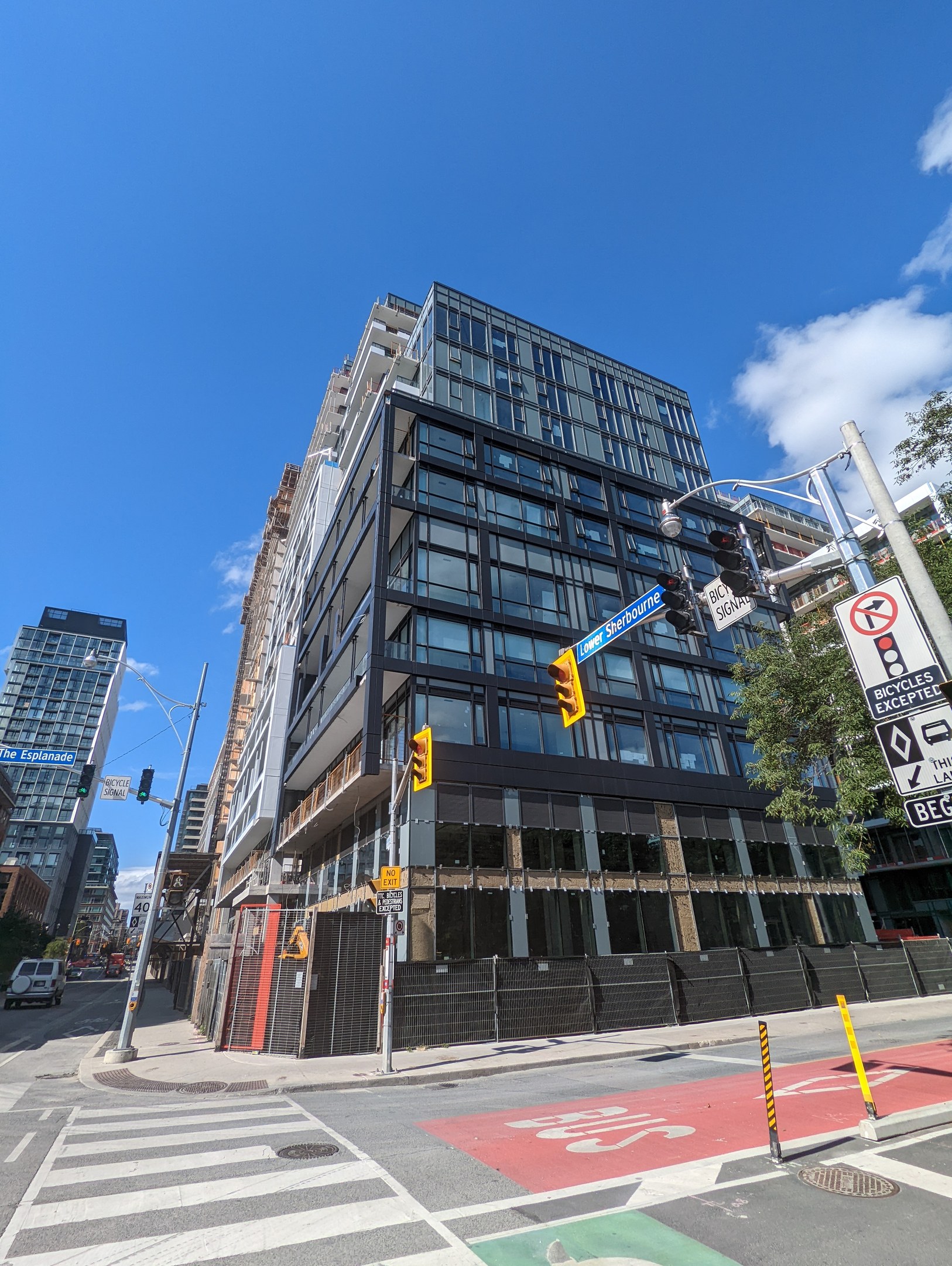 Eine Stadtstraße mit Fahrzeugen, Verkehrsampeln, Schildern, Bäumen und einem Zaun, die zu einem hohen Apartment-/Reihenhausgebäude an 541 East 57th Street, Unit 541 führt, unter einem bewölkten Himmel.