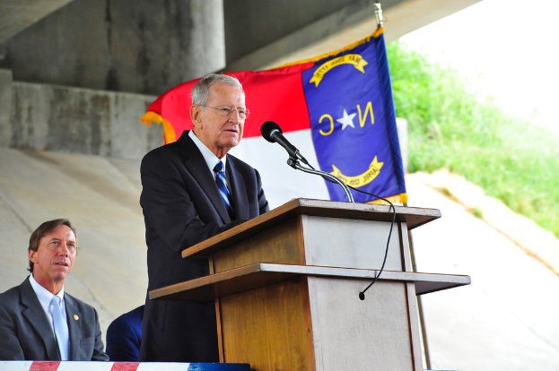 Ein Mann steht vor einem Mikrofon und spricht, im Hintergrund ist eine Flagge zu sehen.