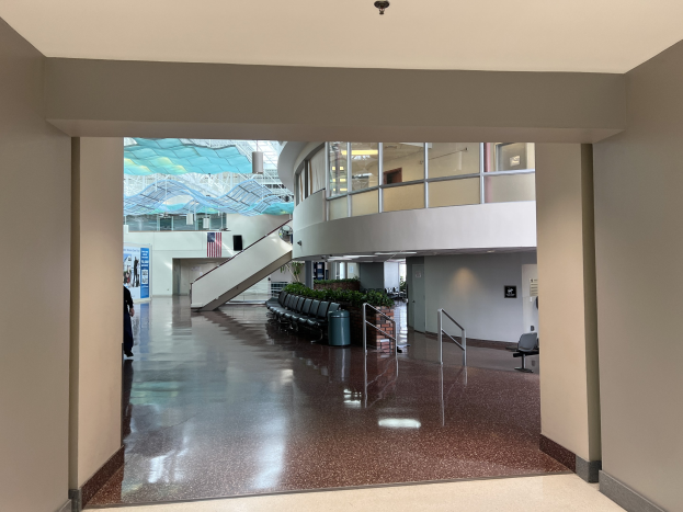 Großer Eingangsbereich mit hohen Decken, zahlreichen Fenstern, einer zentralen Treppe mit Geländern, verstreuten Stühlen, Topfpflanzen, einem Mülleimer, einer stehenden Person, einem Schild und Deckenlampen, die den glänzenden Epoxidharzboden beleuchten.