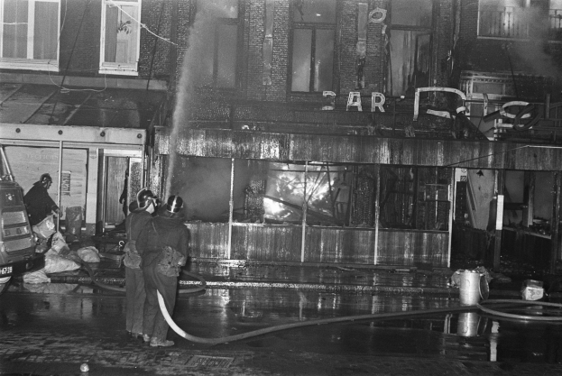 Feuerwehrleute bei der Brandbekämpfung an einer Straßenkreuzung, mit Menschen, die Rohre halten, einem geparkten Fahrzeug, verstreuten Gegenständen, einem Gebäude mit Fenstern, einem Schild und Drähten.