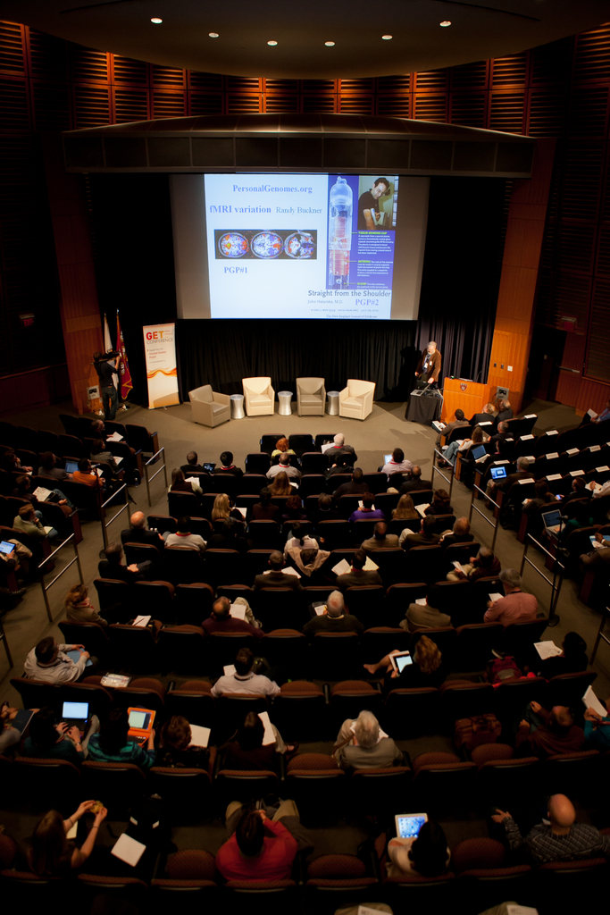 Ein mit sitzenden Menschen gefüllter Hörsaal, beleuchtet von vielen Lichtern, mit Flaggen auf der linken Seite, einem Podium auf der rechten Seite und einer Werbetafel auf der linken Seite.