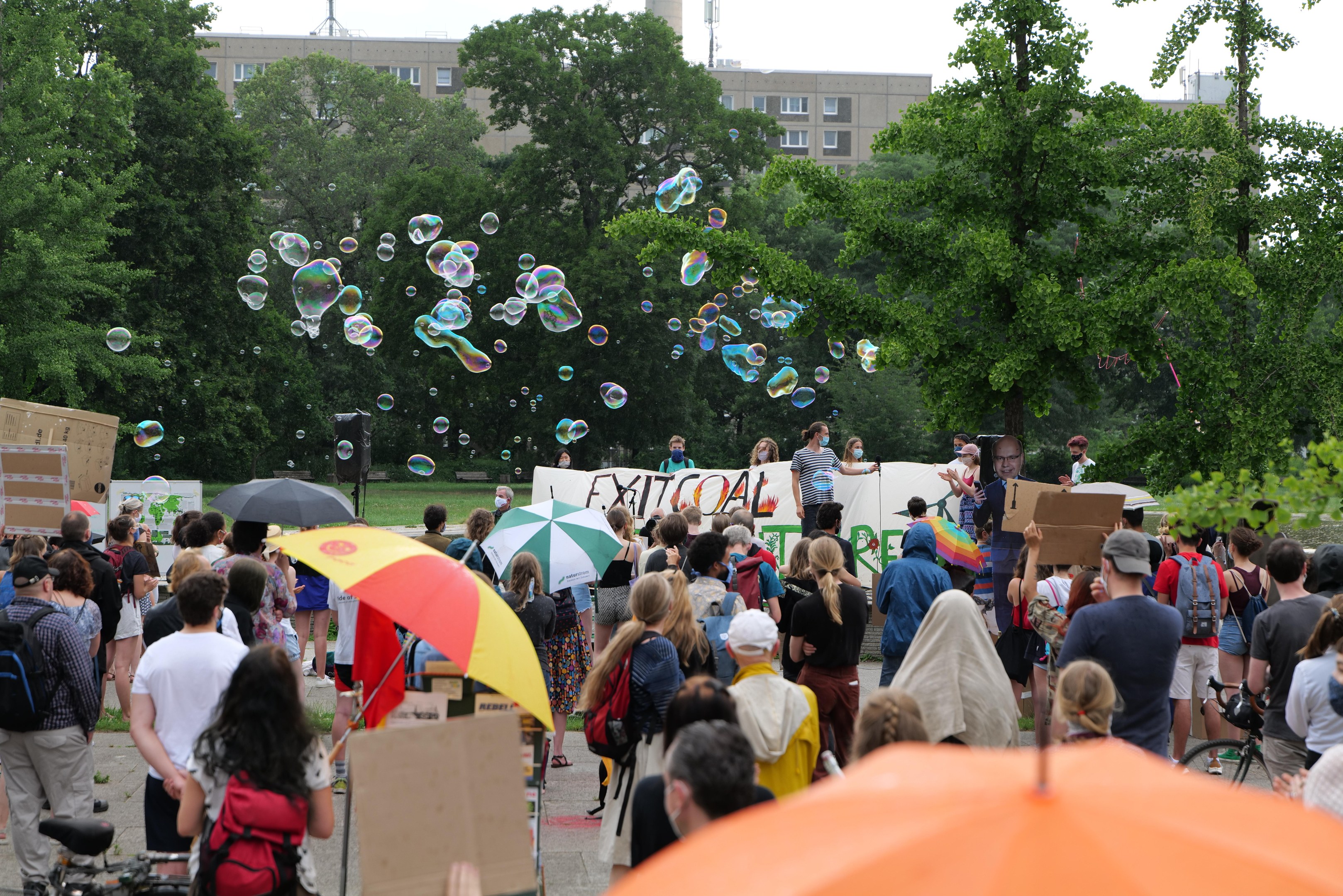 Eine Gruppe von Menschen, die vor einer Menge auf einer Demonstration stehen, einige mit Schirmen, Taschen oder Schildern, andere Seifenblasen pustend, vor einer Kulisse aus Bäumen, Gebäuden, einem Turm und einem klaren blauen Himmel.