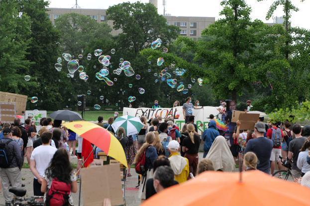 Eine Gruppe von Menschen, die vor einer Menge auf einer Demonstration stehen, einige mit Schirmen, Taschen oder Schildern, andere Seifenblasen pustend, vor einer Kulisse aus Bäumen, Gebäuden, einem Turm und einem klaren blauen Himmel.