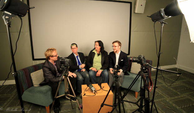Eine Gruppe von drei Personen sitzt auf einem Sofa und gibt ein Interview, wobei eine Person in einem schwarzen Mantel und Hose links, eine Person in einem Mantel und blauer Krawatte mit einem Mikrofon in der Mitte und eine strahlende Person rechts zu sehen sind, mit einem Projektionsschirm dahinter, einem Mikrofonrekorder auf dem Mitteltisch und Kameras mit Stativen auf beiden Seiten.
