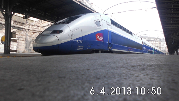 Ein Zug ist in der Mitte des Bildes zu sehen, mit einem Bahnsteig und Text unten. Links ist ein Gebäude mit Dach, Säule und Bahnsteig zu sehen. Oben sind Gebäude, Dächer, Kabel und Himmel zu erkennen.