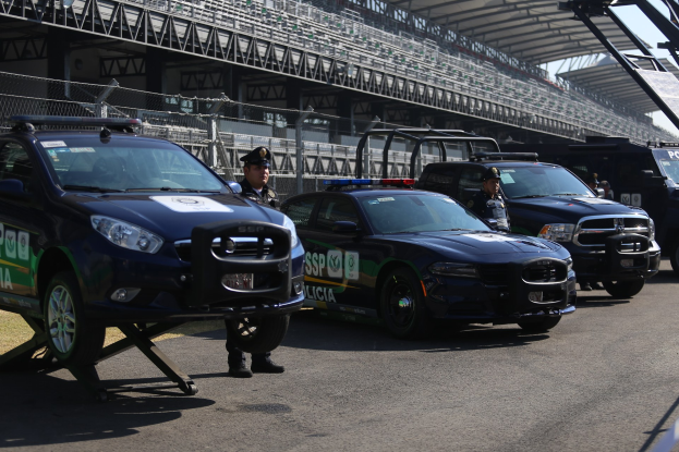 Polizeiautos vor einer Rennstrecke geparkt, uniformierte Personen auf der Straße, Stadion und Himmel im Hintergrund.
