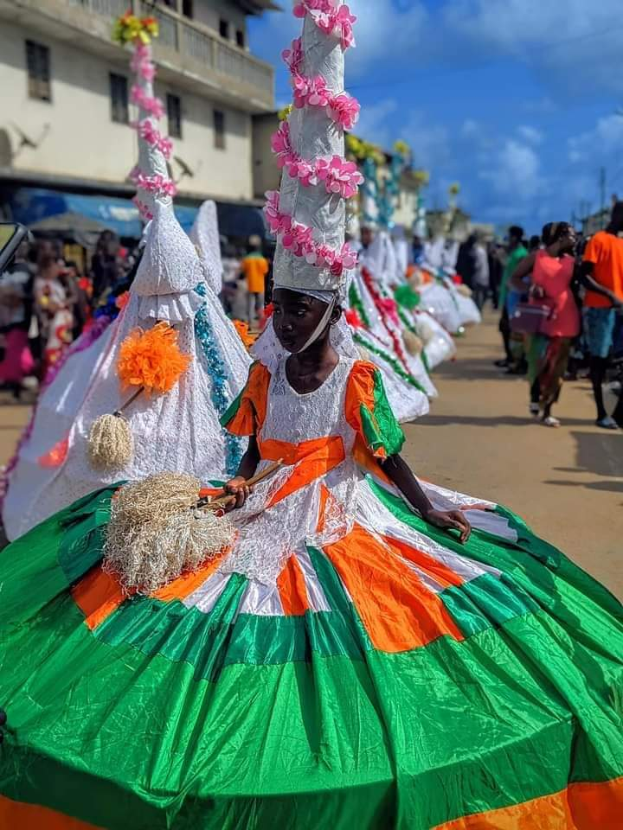 Eine Gruppe von Menschen in bunten Kostümen geht eine Straße entlang, mit einem Gebäude im Hintergrund und einem bewölkten Himmel darüber, wahrscheinlich an einem festlichen Ereignis teilnehmend.