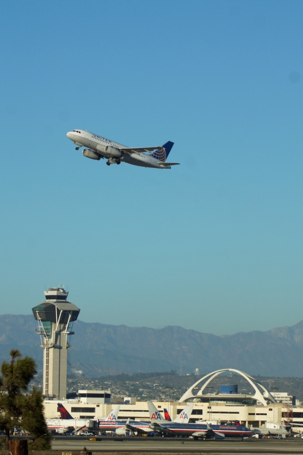 Eine Flughafensicht mit einem weißen Kontrollturm im Vordergrund, geparkten Flugzeugen am Boden, entfernten Gebäuden und einem Flugzeug am Himmel.