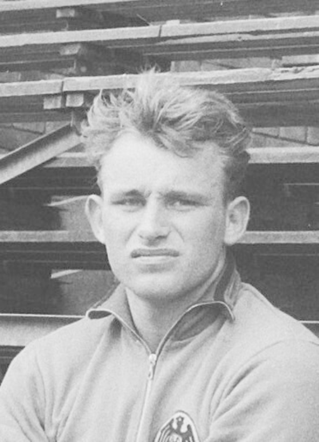 Schwarz-weißes Foto eines Mannes in einer Jacke vor einem Stapel Holzplanken, der als legendärer deutscher Fußballspieler gilt.