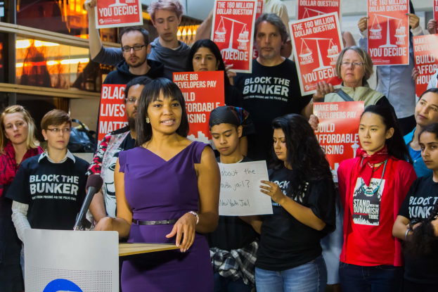 Eine Black-Lives-Matter-Aktivistin spricht in ein Mikrofon an einem Rednerpult, während sie eine Menge anspricht, die Schilder hält, und Bühnenbeleuchtung und Equipment im Hintergrund zu sehen sind.
