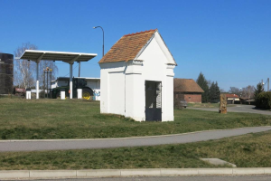 Ein kleines weißes Gebäude mit einem roten Dach, das als erste Tankstelle in Deutschland identifiziert wurde, steht an der Seite einer Straße umgeben von Gras, Bäumen, Polen, Laternen und Häusern sowie einem klaren blauen Himmel.