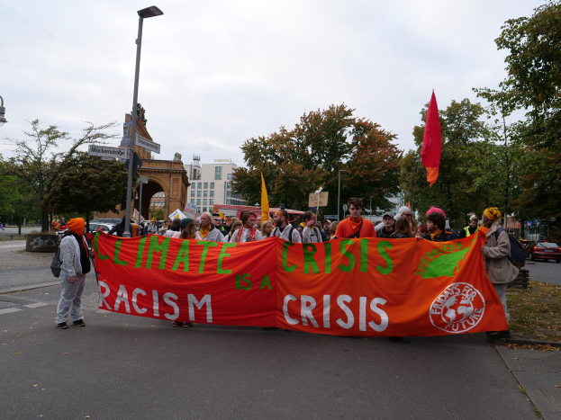 Gruppe von Menschen marschiert durch eine baumbestandene Strasse, haltend ein 'Climate Crisis is a Crisis'-Plakat mit Fahrzeugen und Gebauden im Hintergrund unter einem klaren blauen Himmel.