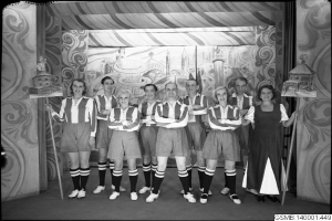 Eine Gruppe lachender Frauen in einem 1940er Fußballteam posiert auf einer Bühne, jede hält einen Stock in der Hand, mit einer bemalten Wand im Hintergrund.