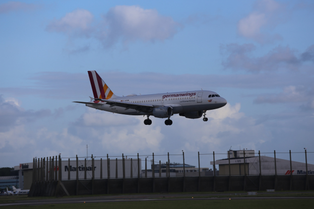 Germanwings Airbus A320-200 beim Abheben vom Frankfurter Airport, mit Gras darunter, einem Zaun im Vordergrund, Gebäuden und Bäumen im Hintergrund und einer bewölkten Himmel