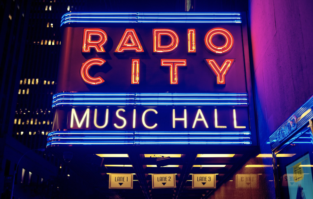 Hell erleuchtetes Radio City Music Hall in New York City mit hohen Gebäuden im Hintergrund, mit einem "Radio City"-Schild und mehreren kleineren Schildern auf der rechten Seite.