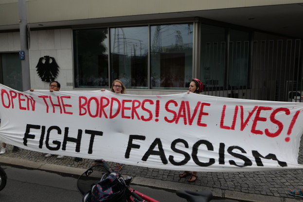 Gruppe von Menschen mit einem Banner mit der Aufschrift "Öffnet die Grenzen, rettet Leben, kämpft gegen Faschismus" vor einem Gebäude mit Glasfenstern und einem Pfahl, mit einem Fahrrad und einer Tasche auf der Straße.