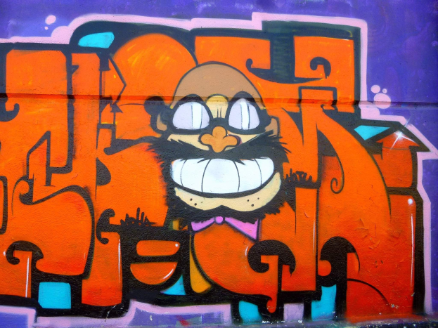 Ein lebendiges Graffiti-Gemälde eines Cartoon-Charakters mit großen Augen, einem breiten Grinsen und einem verschmitzten Ausdruck an der Seite eines Gebäudes, begleitet von auffälligem Text in einer fröhlichen, bunten Stil.