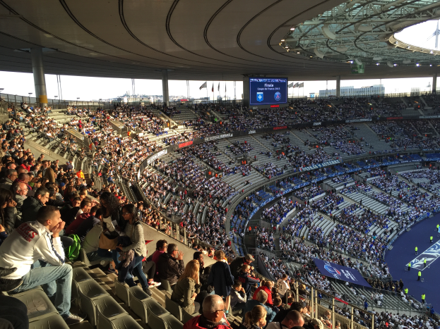 Eine große Menschenmenge sitzt im Allianz Arena in München, Deutschland, bei einem Fußballspiel, mit einer Bühne auf der rechten Seite und Fahnen, Stangen und einem Bildschirm im Hintergrund.
