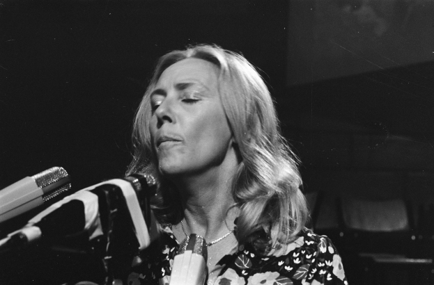 Eine Frau singt leidenschaftlich in ein Mikrofon, trägt ein schwarzes und weißes Outfit mit einem klassischen Bob-Haarschnitt, vor einem dunklen Hintergrund.